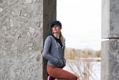 Beautiful young woman is on a run in an urban area while leaning on a concrete wall and looking out at a river while she stops to breath. She is wearing active clothing and a baseball hat.
