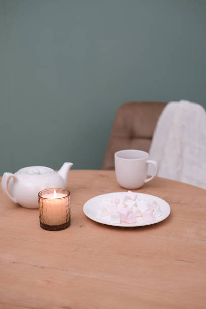 istock Home kitchen interior - white teapot with hot tea, cup of tea, plate with marshmellow, napkin,burning candle, beige chair on green background. 1975398068