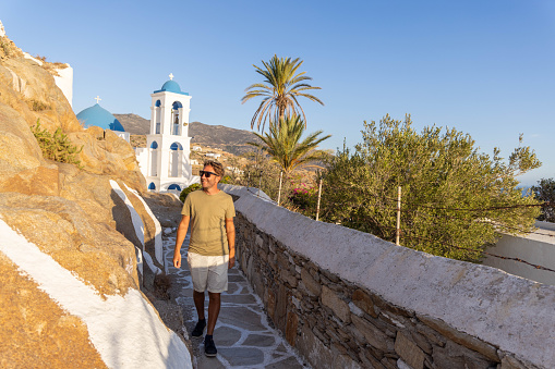 At the golden hour of sunset, a solitary man strolls along a hill overlooking a quaint Greek town. The warm hues of summer paint the sky as he takes in the panoramic view, the serene atmosphere echoing the tranquility of the Mediterranean evening.