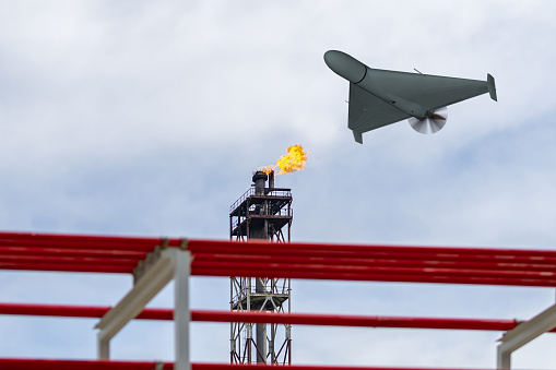 A military drone flies over an oil refinery, fire burning
