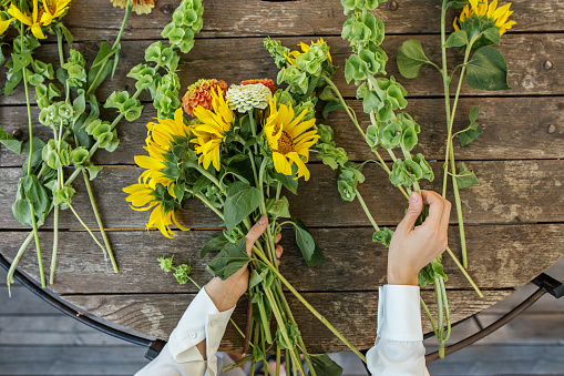 Overhead view of hands arranging a cheerful bouquet of bright sunflowers and orange chrysanthemums on a rustic wooden table.