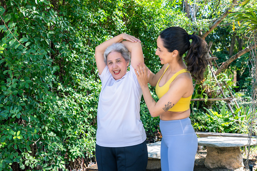 A beautiful Latin mid-adult daughter in sportswear helping and coaching her retirement mother to do stretching and yoga in the home garden, a happy senior enjoying her retirement life