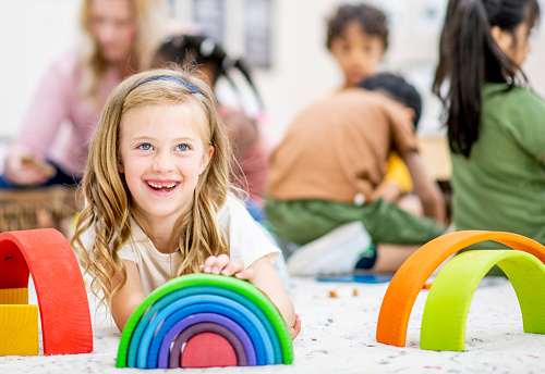 A sweet little blond haired girl lays on the floor of her daycare class room as she stacks rainbow blocks.  She is dressed casually and smiling.