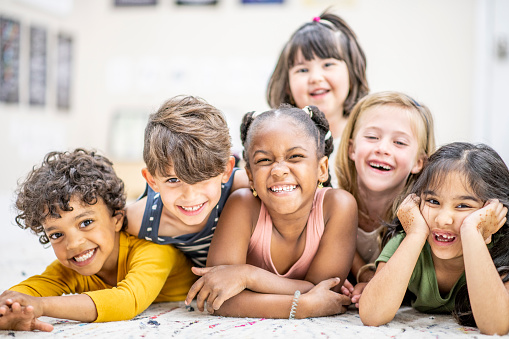 A small group of six daycare friends lay huddled in closely stacked on top of one another as they pose for a portrait.  They are dressed casually and smiling as they enjoy their time together.