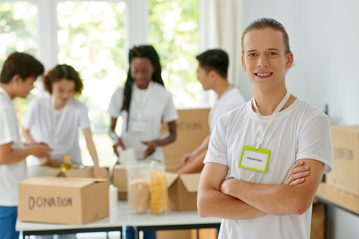 Portrait of smiling volunteer of charity organization, his friend packing donated products in background