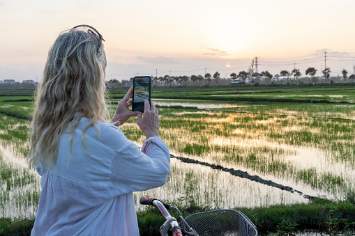 Mature woman pauses bicycle in rice paddies and takes photo