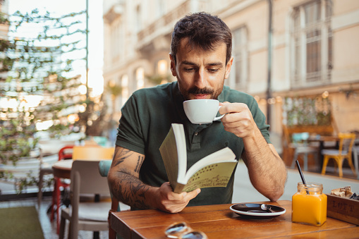 Young bearded guy having coffee break while reading a book in outside cafeteria