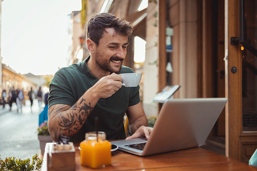 Mid adult handsome man in coffee shop drinking coffee and using laptop in outside cafeteria