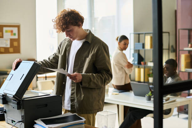 Office manager with financial papers Young office manager in casualwear looking at financial paper while standing in front of xerox machine in coworking space computer printer stock pictures, royalty-free photos & images