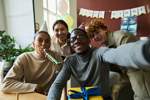 Group of happy young intercultural office workers in birthday caps looking at camera while posing for selfie during office celebration