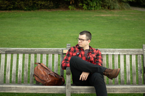 young man with punk style waiting on a park bench with a brown leather backpack and red flannel jacket. Green background.