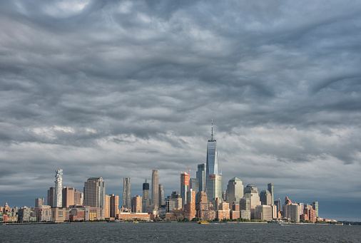 One World Trade Center dominates the Lower Manhattan skyline in this view from Hoboken, New Jersey. Late afternoon light creates a spotlight effect on the buildings as the eastern sky is overcast with dramatic clouds.