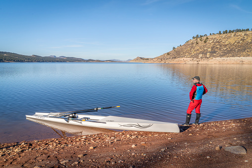 Loveland, CO, USA - January 30, 2024:  Senior male rower and a coastal rowing shell by Liteboat is landing on a rocky shore of Carter Lake in fall or winter scenery in northern Colorado.