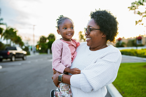 An African American woman carries her three year old daughter on her hip as they walk around their neighborhood in Florida at sunset.