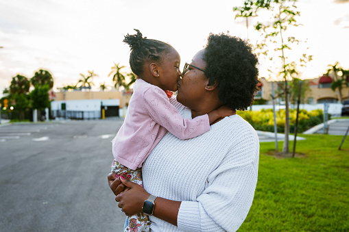 An African American woman carries her three year old daughter on her hip as they walk around their neighborhood in Florida at sunset, and gives the child a kiss.