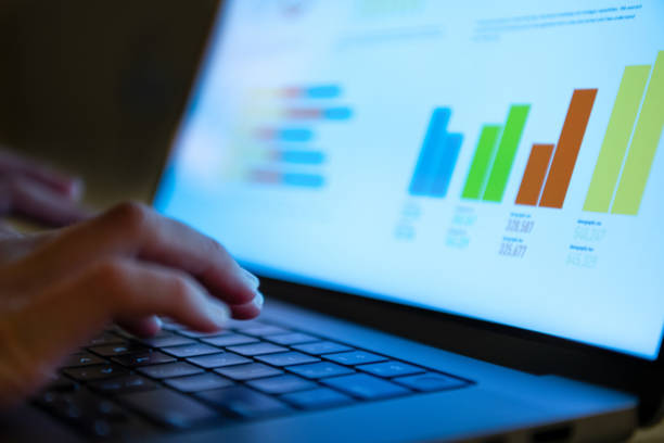 Close up of a woman’s hands typing on a laptop computer keyboard. There are finance charts and graphs on the device screen.