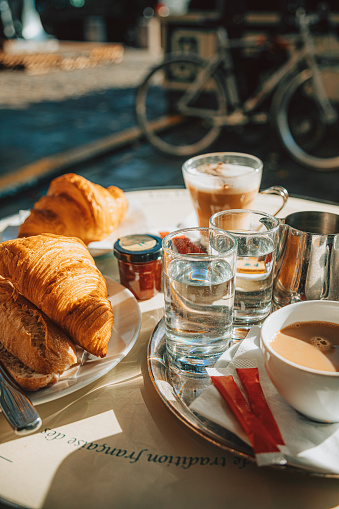 A delightful tableau unfolds in a Parisian street cafe on a sunny morning, where a close-up captures the quintessential pairing of a freshly brewed cup of coffee latte and a buttery croissant. In the background, the soft morning sunlight casts a warm glow, illuminating a spread of orange juice, tea, and a baguette sandwich with butter and jam. This scene evokes the essence of a leisurely start to a beautiful day in the City of Light