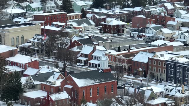 American flag in Small Town USA covered in winter snow. Church and colorful business buildings at town square in Manheim Pennsylvania USA. Aerial long zoom.