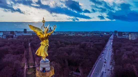 Drone point of view on Victory column  (67m, built 1864) with observation desk  and Roundabout on Grosser Stein square: 
Amazing sunbeams of sunset,  winter forest of Tiergarten.   Winter the Tiergartenwith naked trees.  Cars are moving. 
  Soon  Christmas in Berlin,  German capital.