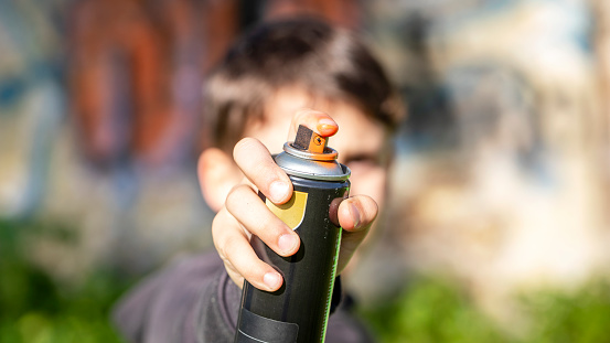 Child painting with color spray pointing at the camera with the background out of focus. Detail of the aerosol valve or nozzle in close-up held by a child on the street.