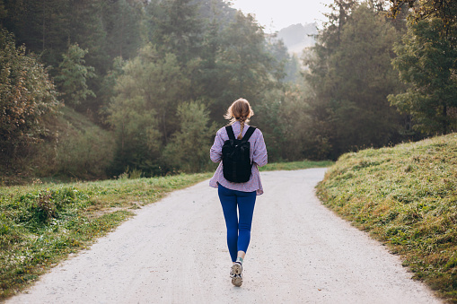 Young female with backpack hiking on the road in nature. Beautiful 30s woman walking in forest in running clothes. Wanderlust travel concept, atmospheric moment