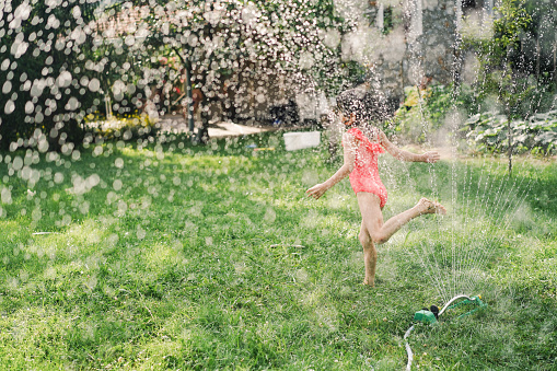 Little girl having fun jumping over garden sprinkler and refresh herselves on a hot summer day