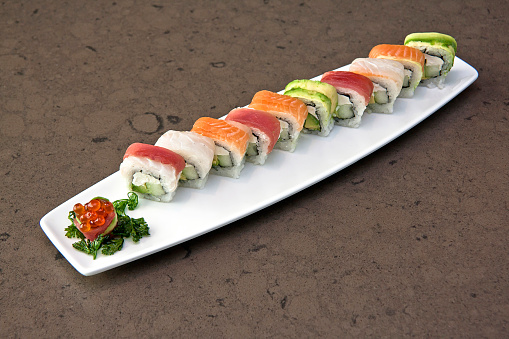 Assortment of Japanese sushi rolls nigiri, sashimi, maki, salmon, tuna and avocado served in long rectangular plate over stone background