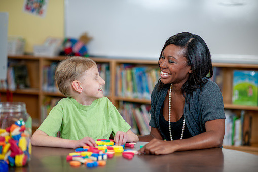 An Occupational Therapist works with a young boy in a wheelchair with the help of multicoloured blocks.  She has laid out various shapes on the table and is working on the boys dexterity with the use of the blocks.