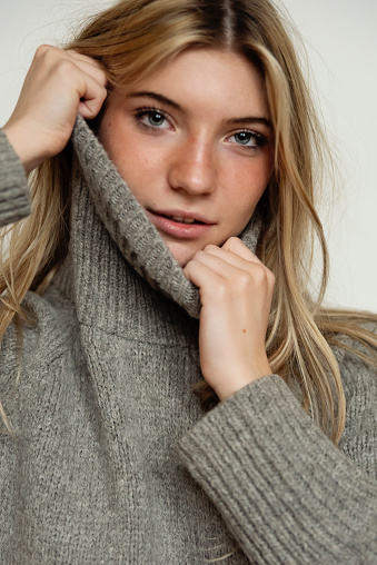 Portrait of beautiful young woman of generation Z on white background. She has long blond hair and is wearing a grey wool turtleneck. She is playing with her sweater, smiling and looking at camera. Vertical head and shoulder studio shot with copy space. This was taken in Montreal, Quebec, Canada.