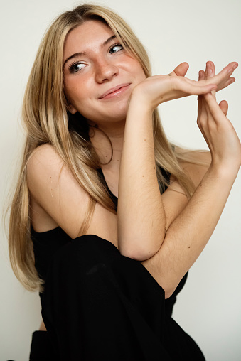 Portrait of beautiful young woman of generation Z on white background. She has long blond hair and is wearing a black tank top and pants. She is sitting on the ground. Vertical waist up studio shot with copy space. This was taken in Montreal, Quebec, Canada.
