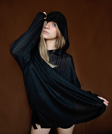 Portrait of beautiful young woman of generation Z on deep brown background. She has long blond hair and is wearing a striped black hoodie over black cropped top and shorts. She is standing, obscuring her face. Vertical mid length studio shot with copy space. This was taken in Montreal, Quebec, Canada.