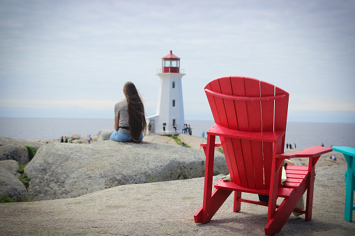 Peggy's Cove, Nova Scotia, Canada - August, 2023: Red Adirondack chair in front of the Peggy's Point Lighthouse in the historic fishing town of Peggy’s Cove, Nova Scotia, Canada, during a summer day in August 2023.