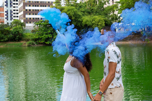 Happy couple discovering that their baby is a boy through the blue smoke. Gender reveal. Against green colored lagoon.