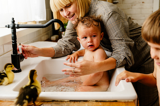 Baby boy  is playing with duck birds while his mother bathing him in the kitchen sink