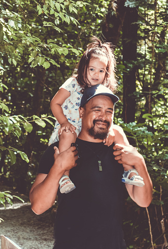 A little girl rides on her father's shoulders walking through an outdoor forest park.