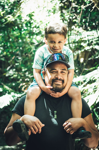 a young Maori boy riding on top of his Dad's shoulders outdoors in a park.