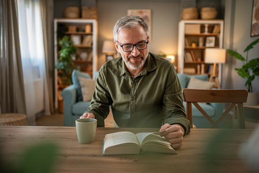A mature man enjoys a quiet moment reading a book, sitting at a table with a cup of coffee in front of him