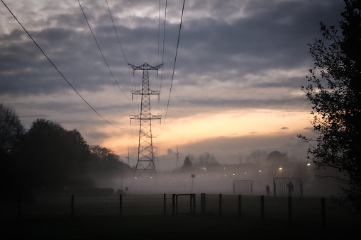 Foggy sunset over a playfield in a residential area of ‘s-Hertogenbosch (den bosch), located in the province of North Brabant. In the foreground is a transmission tower and all the way in the background are some windturbines. Also, there’s 2 people in silhouette playing soccer.