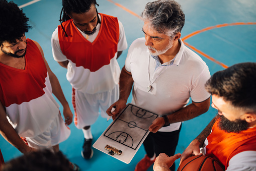 From above view of a senior basketball trainer working on game tactic with his multicultural team on court. An interracial basketball players standing on court with trainer, working on game strategy.