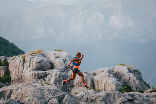 An extreme sportswoman is skyrunning and jumping on rocky mountain while trail running and mountaineering. A fit runner is doing her training and challenging herself on height. A sportswoman adventure