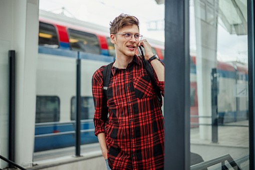 Handsome man commuting to work by a train while talking on a smartphone. Hipster guy is talking on his phone while waiting for a train to arrive at the railway station. People travel transport concept