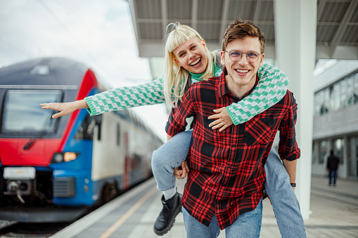 Portrait of urban travelers having piggyback ride at train station.A young couple is acting silly and having piggyback ride outdoors. There is a train on railway in a blurry background. Travel concept