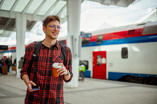 Portrait of a happy commuter walking on a train station with coffee to go and a phone in his hands. A young passenger is smiling and walking on a railway station. There is a train in a background.