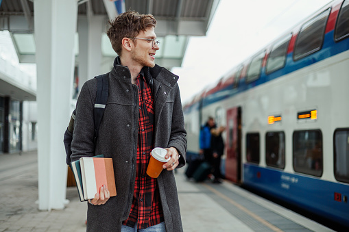 A young urban man is sanding at train station and waiting for a train. Traveler is standing at railroad station with books in his hands and looking at the train. A passenger is preparing for a trip.