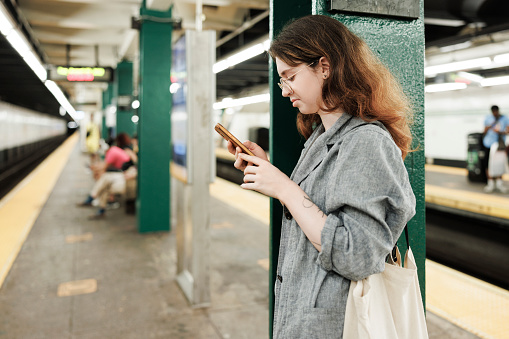 Young woman in eyeglasses using telephone on a platform of subway train station on blurred underground view
