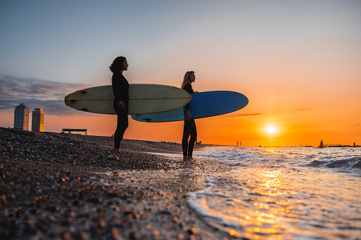 Two surfer girls admiring the amazing view of the sea and waves before going for an early morning surfing session. The sun is rising and the sky is orange and blue. The female surfers are carrying their surfboards and are wearing black wetsuits.