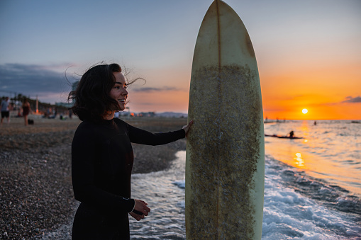 An attractive young adult female surfer standing on a beach next to her surfboard. She is preparing for a relaxing early morning surfing session. The sky is orange due to the sunrise. The woman looks happy and calm. She is wearing a wetsuit.