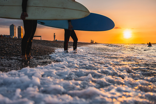 Close-up of two female surfers stepping into the water before a morning surf session during sunrise. Only legs and surfboards are visible, no face. The sun is rising and the sky is orange. Idyllic view of the beach and horizon. Beautiful surfing conditions.