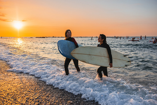 Happy female surfers smiling while walking out of the water during sunrise. They are wearing wetsuits and are carrying their large surfboards.