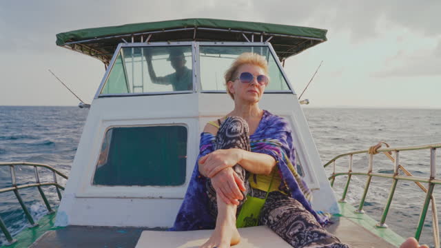 A 50-years-old active European woman, a tourist, is resting on the deck of a small fishing boat during the deep sea fishing trip in Sri Lanka when a captain is ruling the ship from a deckhouse in the backdrop.